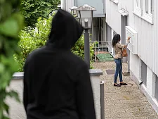 Vermummte, dunkel gekleidete Person beobachtet aus der Ferne eine Frau, die an einem Postkasten steht 