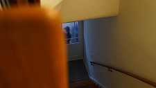 Ein Treppenaufgang, an dessen Ende eine vermummte, dunkel gekleidete Gestalt hinter einer Tür zu erkennen ist.